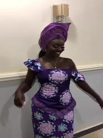 A young, dark-skinned woman stands in front of a light-colored wall, wearing a matching purple dress and headwrap. The dress has an off-the-shoulder design with ruffled edges, and the skirt is a similar pattern to the top. The headwrap is also purple, wrapped high on her head. She is smiling and posing, occasionally turning her head to show different angles. She has a necklace on and a watch on her wrist. The wall behind her has a white trim and a light fixture.