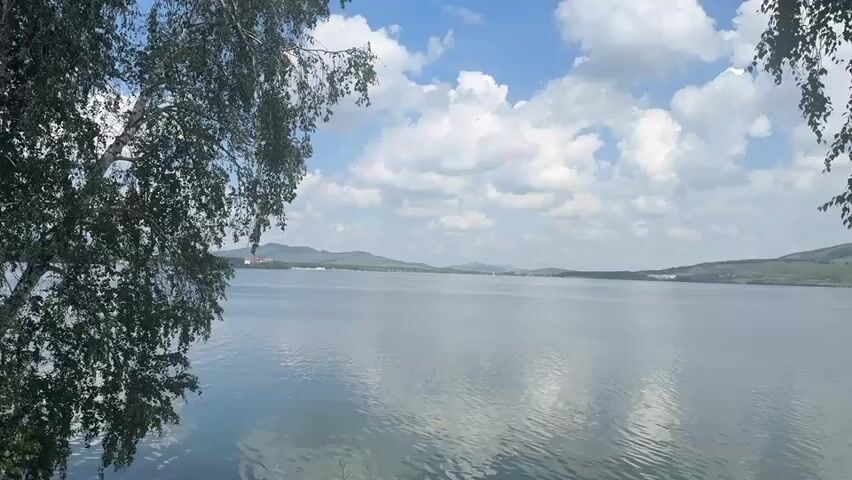 This media file depicts a serene natural landscape. The scene is dominated by a large, calm body of water, likely a lake, reflecting the sky. In the foreground, on the left and right sides of the frame, are the overhanging branches of trees, their leaves a vibrant green, framing the view. Beyond the water, the distant horizon is marked by rolling hills or low mountains, covered in greenery. The sky above is a bright blue, dotted with numerous fluffy white clouds, suggesting fair weather. The lighting indicates it is daytime, likely mid-morning or afternoon.

There are no people or distinct objects visible in the scene, emphasizing the natural beauty and tranquility of the location. The activity taking place is simply the passage of time, with the gentle ripples on the water and the movement of clouds creating a sense of peaceful stillness. The overall emotion evoked by the imagery is one of calm, relaxation, and appreciation for nature. The location context of Yakty-Kul, Russia, places this scene in a region known for its natural beauty, often featuring lakes and hills. No text is visible in the media file.