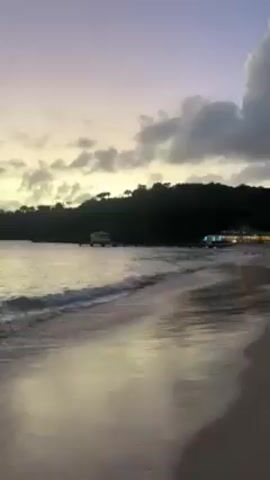 The video shows a beach at dusk. The sky is a mix of purple, yellow, and gray.  A dark, tree-covered hill forms a backdrop.  Two small buildings are visible in the distance, one a simple structure on stilts over the water, and the other larger with lights. Gentle waves roll onto the sandy beach.  The wet sand reflects the colors of the sky. No people are visible.