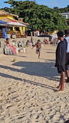 On a sunny, sandy beach, a young boy in shorts is attempting to kick a coconut.  He runs and kicks it, then falls. The boy sits for a moment, then gets up and walks away. The beach also features other people, beach umbrellas, chairs and assorted beach equipment. There is a yellow building in the background.