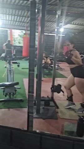 A woman in a black sports bra and black shorts poses in front of a mirror in a gym while taking a selfie with a pink phone. She wears ankle weights and gray sneakers. The gym has green turf flooring and various weight machines, including a squat rack behind her. Other people can be seen working out in the background. The gym appears to be well-lit with exposed metal beams visible on the ceiling.
