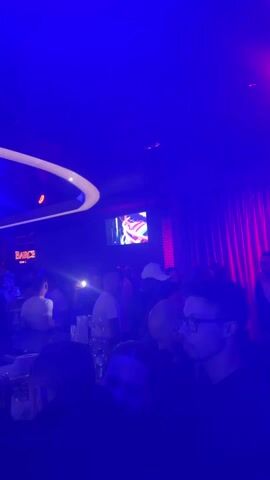 The video shows the interior of a crowded bar or nightclub.  The lighting is primarily blue, with red accents.  People are standing and talking, seemingly enjoying themselves.  A bar runs along the side of the room. There are bar stools lining the bar. There are flat screen TVs hanging on the walls, displaying colorful, abstract patterns.  A sign that reads "BARCI" is also visible. The ceiling has modern, curved lighting fixtures. The video seems to have been shot while walking through the crowd.