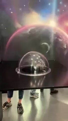 The video shows an adult with glasses popping their head up into a clear dome on a table. The dome has Christmas-style lights hanging inside of it. The table is black and there is a colorful planetary background on the wall behind it. There are two other people whose legs are visible. The floor is a glossy gray.