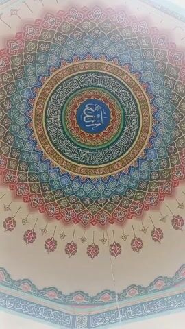 The video shows a low angle view of a mosque's dome. The dome has intricate, circular patterns in shades of blue, green, pink, and brown.  Arabic script is prominently featured in the center, surrounded by concentric circles. There are repeating floral motifs around the edges of the dome.