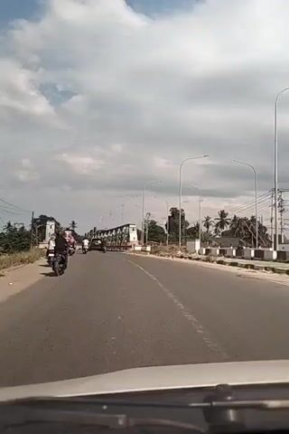 The video shows a car driving towards an old steel truss bridge on a two-lane road. There are motorcycles on the road ahead of the car and streetlights along the roadside. Trees and power lines line the road in the background. The sky is blue with some white clouds.