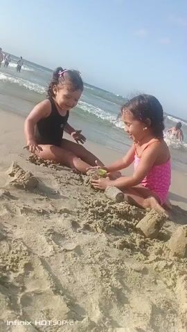 Two young girls are playing on a sandy beach. One girl, wearing a black swimsuit, is sitting on the sand. The other girl, in a pink swimsuit, is also sitting and appears to be interacting with the first girl and a green sand mold. In the background, the ocean waves are visible, and there are a few other people in the water and on the beach. The scene suggests a sunny day at the beach, likely during daylight hours given the brightness of the sky. The children's body language suggests engagement and play. The visible text at the bottom of the frame reads "Infinix HOT 50Pro." The context provided indicates the location is Naiguatá, Venezuela.