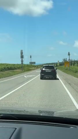 A black SUV is driving on a highway, viewed from inside another vehicle. The highway is paved and has white lane markings. On the right side of the road, there are signs, including a yellow sign with Russian text that appears to be a warning, and a circular sign with the number 70, indicating a speed limit. The surrounding landscape consists of green fields, possibly vineyards, and trees. The sky is blue with scattered white clouds. The video shows the SUV from behind as it moves away from the camera.
