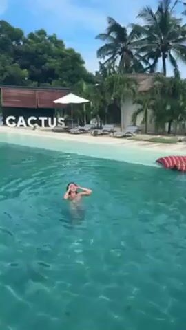 The video shows a woman swimming in a pool. She floats on her back, moving her arms. The water is turquoise. Behind her, the word "CACTUS" is displayed in large white letters, standing on a white surface that resembles a beach. There are lounge chairs, a white umbrella, and palm trees in the background. The sky is blue with some clouds.