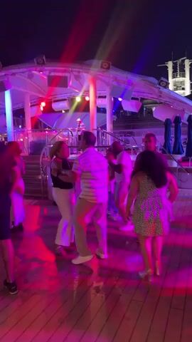 The video shows a group of people dancing on a wooden deck at night. There are bright pink and purple lights illuminating the scene, with some red and blue spotlights shining from above. In the background, there is a structure with lights and railings, possibly part of a ship or a venue with multiple levels. Several couples are dancing, some in close embrace, while others dance individually. The people are dressed casually, with some wearing shorts and t-shirts, and others in dresses or more formal attire. The overall atmosphere appears to be lively and celebratory.