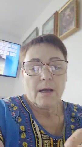 A middle-aged woman with short, light brown hair and rectangular, reddish-brown glasses is captured in a close-up, self-shot video or photograph. She wears a vibrant blue top featuring a multi-colored, intricate pattern with geometric shapes and stylized figures in yellow, red, black, and white. Her mouth is slightly open, suggesting she is speaking or reacting, and her right index finger is pressed against her lower lip, possibly indicating thought or contemplation.

The setting appears to be an indoor room, likely a home, with plain white walls. In the background to her left, a flat-screen television is visible, displaying reversed text which appears to be a commercial or brand advertisement. The text "O'KEEFFE'S" is clearly legible on a red background, followed by what looks like "GUARANTEED RELIEF." To her right, on the white wall, two framed pictures are hanging; the one closer to the camera features a portrait in a gold-colored frame. The room is brightly lit, suggesting it is daytime or well-lit artificially.