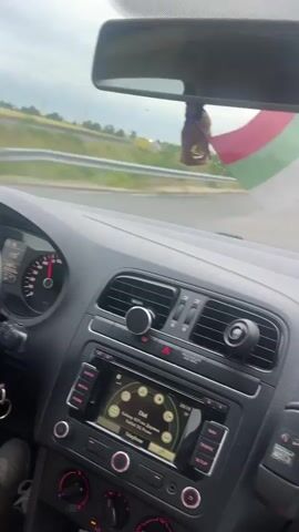 The video shows the interior of a car driving on a road. A Bulgarian flag is hanging from the rearview mirror. The car's infotainment system is visible, displaying navigation information. The driver's hands are not visible. The road appears to be a highway with guardrails and signs. The sky is overcast. The overall impression is a journey through a rural or suburban landscape.