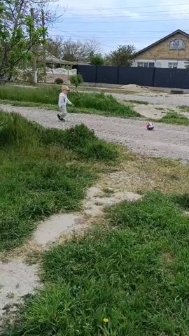 A young child wearing a white jacket, tan pants, and a tan cap walks on a dirt road and then kicks a red and white ball. The road is surrounded by green grass and weeds, with a few patches of bare dirt. Behind the road is a gray fence and a house with a gable roof. There is also a smaller building with a white picket fence in the distance. The sky is partly cloudy.