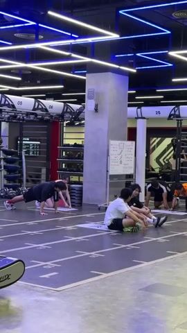 This is a short, static video clip of a gym. In the foreground, a portion of a black and silver speedfit treadmill is visible on the left. The main focus is on the middle ground where several individuals are engaged in fitness activities. Two people are in the foreground, seated on the floor with their legs extended, facing away from the camera. Behind them, further to the right, a few more people are in various workout poses on mats. On the left side of the middle ground, two people, a male and a female, are performing a plank exercise. The male is in a standard plank position, while the female is behind him in a modified plank, resting on her knees. In the background, gym equipment is visible, including racks of weights and what appear to be exercise machines. The gym features a modern industrial design with exposed ceilings, blue LED strip lighting, and concrete walls. The floor is marked with lines and numbers, suggesting designated workout areas. A whiteboard with writing is mounted on a pillar in the middle ground. The overall ambiance is that of a busy, contemporary fitness facility.