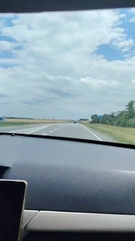 A first-person view from inside a car shows a highway stretching into the distance under a partly cloudy sky. The dashboard and a phone are visible in the foreground. Cars can be seen on the road ahead, and there are signs and trees lining the sides of the highway. The scene suggests travel on a sunny day.