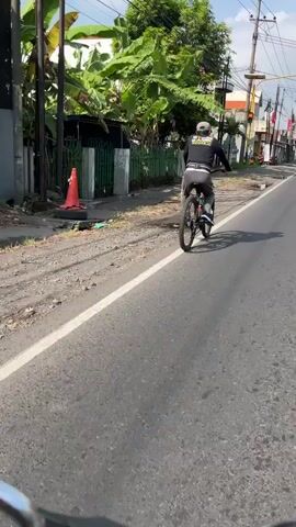The video shows a person riding a bicycle on a paved road in what appears to be Siring, East Java. The cyclist, wearing a black long-sleeved shirt, gray pants, and a cap, is seen from behind, riding away from the camera. The bicycle is a mountain bike with knobby tires.

The scene is set outdoors during the daytime, with bright sunlight casting shadows. The weather appears to be warm and sunny. To the left of the road, there is a sidewalk with a green fence and lush vegetation, including large banana leaves and trees. Behind the fence, a building with a white and brown facade is partially visible. Further down the road, more buildings and power lines can be seen, suggesting an urban or semi-urban environment.

The road is asphalt with a white line marking the edge. The overall atmosphere seems casual and everyday, with no indication of any special event. The cyclist appears to be casually riding, possibly commuting or exercising.

Visible text on the cyclist's shirt reads "STOP SEDIH" which translates to "STOP SAD".
