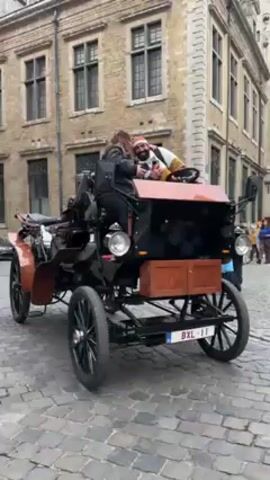 A video shows a vintage car with two passengers sitting in it. The car is black with dark brown wooden details and has a license plate reading BXL-11. It is parked on a cobblestone street in front of buildings with old European architecture. The person driving is wearing a white sweatshirt with brown and orange stripes and a red hat. They are gesturing with their hands and speaking to the passenger. The passenger is wearing a black jacket. The camera circles around the car, showing the building and other people walking by.