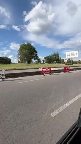 This media file depicts a street scene in Dnipro, Ukraine, on a sunny day with scattered clouds. The foreground shows a paved road with some damage or construction, indicated by excavated areas and temporary barriers. Red and white construction barriers are strategically placed to redirect traffic and alert drivers to the work zone.

In the mid-ground, there's a grassy area with trees, suggesting a park or a landscaped median. A large billboard is prominently visible, advertising "REVO ENERGY" with an image of what appears to be a cartoon character or mascot. Further in the background, more trees and some distant structures can be seen, hinting at an urban or suburban environment. A statue or monument is also discernible in the distance, adding a point of cultural interest to the landscape.

The overall atmosphere is one of ongoing infrastructure work, typical for a city. The weather appears pleasant, with a bright blue sky and fluffy white clouds contributing to a feeling of a warm day. The presence of the billboard suggests a commercial area. No people are clearly identifiable or engaged in specific actions within the frame, making the focus primarily on the environment and the construction activity. The time of day is likely daytime, given the bright sunlight casting shadows. The combination of road construction, greenery, and a prominent advertisement creates a dynamic urban landscape.