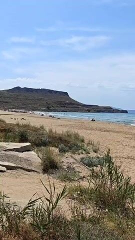 The video captures a serene daytime scene on a sandy beach, likely in Gouves Municipal Unit, Greece. The sky is a bright blue with scattered white clouds, indicating fair weather. In the foreground, dry grass and low-lying bushes grow in the sand. Beyond this vegetation, a wide expanse of light brown sand stretches towards the clear, calm ocean, where gentle waves can be seen breaking on the shore.

Several people are present on the beach and in the water. Some are swimming or wading, while others are relaxing on the sand, with a few beach umbrellas (one orange, one blue, and one green) providing shade. At the 0:02 mark, a person is prominently featured walking across the sand, carrying a large, folded blue inflatable raft, presumably heading towards the water.

In the distance, across the blue water, a long stretch of land, possibly a large island or part of the mainland, with hills or mountains, is visible under the hazy sky. To the left, rugged coastal hills form the backdrop of the beach. The overall impression is one of a peaceful and enjoyable day at the seaside. No discernible text is visible in the video.
