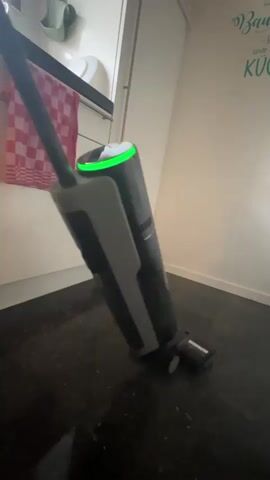 A handheld wet dry vacuum cleaner is shown in a kitchen setting. The vacuum is dark gray and black with a glowing green ring around its display. A person's hand is visible at the top of the vacuum's handle, operating it. The vacuum is positioned on a dark tiled floor that appears wet in places. Behind the vacuum, a white kitchen cabinet with a red and white checkered towel hanging from a handle is visible on the left. The wall behind the vacuum has green text written on it. The vacuum is tilted slightly to the left.