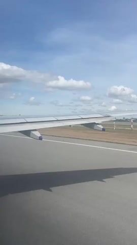 The view from inside an airplane shows the wing of the plane and the runway during takeoff. The sky is visible with scattered clouds. Beyond the runway, there are buildings, trees, and fields. The plane is moving forward on the runway.
