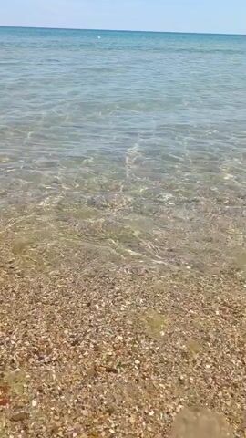 A video shows a shallow body of water at the edge of a beach. The water is clear and calm, with small waves gently lapping at the shore. The seabed is covered in small pebbles and shells. The sky is clear and blue.