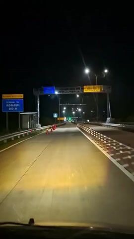 The video captures a first-person perspective from inside a moving vehicle driving on a highway at night. The road is illuminated by the car's headlights and overhead streetlights. There are multiple lanes, and the vehicle appears to be in the leftmost lane. On the left side of the road, there's a barrier with orange traffic cones placed along it. Further down the road, there are large overhead gantry signs with blue and yellow panels, some with text. In the distance, more streetlights are visible, creating a tunnel-like effect of light. The sky is dark.