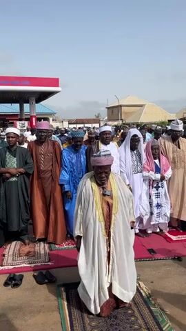 A crowd of men and one small child stand on prayer rugs to pray outdoors. They are standing in rows, and most are wearing traditional Muslim garb. Some of the men are wearing hats. They appear to be praying in front of a gas station in a residential area. The weather is sunny and clear.