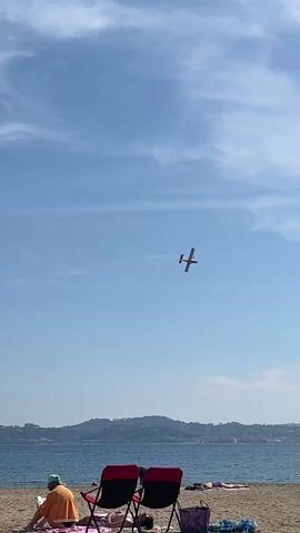 The video shows a small plane in the sky above a beach. The sky is mostly clear with some scattered clouds. In the foreground, there are two beach chairs with people sitting in them, and other people lying on the beach. The beach is sandy, and there is water in the background. A line of green hills can be seen in the distance across the water. The plane is flying at a distance and seems to be moving.