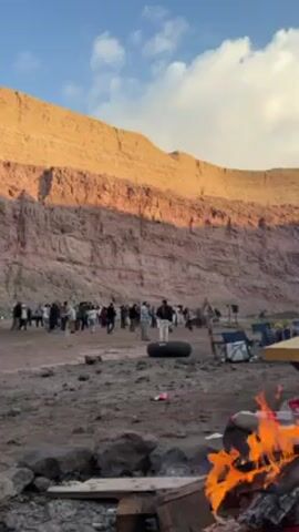 The video shows a group of people gathered in a dusty, rocky area near a large red rock formation. In the foreground, a campfire blazes, casting light on the immediate surroundings. The group is composed of a diverse mix of people, some standing and others moving around. There's a tire placed in the middle of the group. The background is dominated by the imposing rock formation, which is illuminated by the late afternoon sun. The sky is blue with some white clouds. The camera remains relatively still. The overall impression is that of a social gathering or a campsite set against a dramatic natural backdrop.