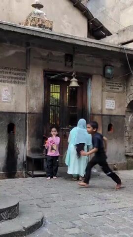 A group of people are gathered in an outdoor courtyard in front of a building. The building appears to be a temple, with a large wooden door in the center and inscriptions on the walls. A bell hangs above the door.

In the foreground, a young boy wearing a dark t-shirt and black pants is running and appears to be playing. To his left, a young girl in a pink t-shirt and black pants stands and watches.

In the center of the frame, a woman wearing a light blue traditional Indian outfit holds a baby. She lifts the baby up towards the bell above the temple door. Another woman, dressed in an orange traditional Indian outfit, stands to the right, facing the temple and appears to be praying with her hands clasped.

The courtyard is paved with stone, and there are steps in the foreground. The lighting suggests it is daytime.