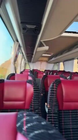 The video shows the interior of an empty bus. The seats are upholstered in red leather with a plaid pattern of black, red, and white. The bus appears to be in motion, as indicated by the blur of the scenery outside the windows. The ceiling of the bus has overhead compartments and lighting fixtures. There are also windows along the side of the bus, some of which are covered by white curtains. The overall impression is one of a comfortable and modern bus interior.