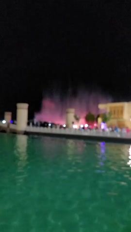 The video shows a nighttime view of a body of water, likely a canal or large pond, with a promenade on the far side. The water is a vibrant green and reflects the lights from the scene. On the promenade, there are several tall, cylindrical structures that appear to be part of a building or attraction. Behind these structures, a water fountain display is in operation, with plumes of water illuminated by colorful lights, predominantly pink and white. There are numerous people visible on the promenade, some walking and others gathered to watch the fountain. The background is a dark night sky. The overall scene suggests a lively public space with a water feature attraction at night.