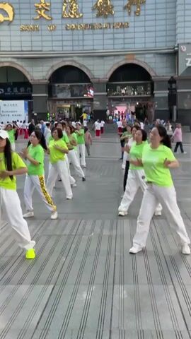 A group of women are dancing in front of a building that reads "BANK OF COMMUNICATIONS". The women are mostly wearing matching neon green t-shirts and white pants. One woman has a white baseball cap. They are arranged in rows and performing synchronized dance moves. The location seems to be a public square with people walking around in the background. The day seems bright and sunny.