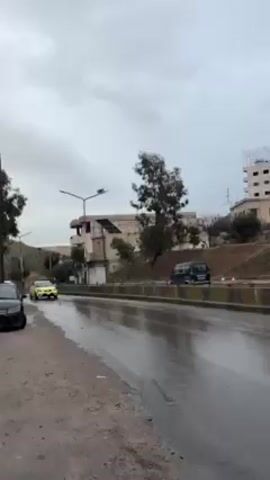 The video depicts a wet, paved road on a cloudy day. A lime-green car drives on the road. There are buildings and trees in the background. A dark minivan is parked further down the road. Several streetlights are visible. Another car is parked to the left of the frame.