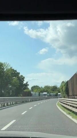 The video shows a point-of-view shot from inside a vehicle traveling on a highway on a sunny day with scattered clouds. The road is paved with white lines marking the lanes. There are guardrails on both sides of the road. Trees and sound barriers line the sides of the highway. Several trucks are visible on the road. A pedestrian overpass is seen in the distance. The audio includes a voice stating "85 kilometers, 9:20."