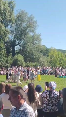 A group of children are playing a game of tug-of-war on a sunny day in a grassy field. A man in a yellow shirt and yellow pants is officiating the game. A crowd of spectators is watching the game. Trees and hills are visible in the background.
