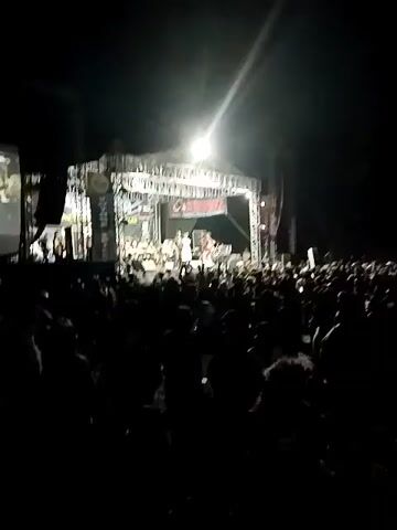 A concert is taking place at night. A large crowd of people are gathered in front of a brightly lit stage. On the stage, a band is performing. The stage is set up with multiple speakers and lighting equipment. A large banner hangs behind the band with text that is difficult to read due to the lighting. The crowd appears to be enjoying the performance, with many people raising their hands.