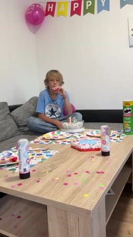 A teenage boy is sitting on a couch with a party blower to his mouth, and a birthday cake with candles shaped like the number 13 in front of him. The cake is on a table covered in confetti and party supplies. A pink balloon and a "Happy Birthday" banner are visible in the background. The boy is wearing a blue t-shirt and jeans. The environment appears to be a living room.