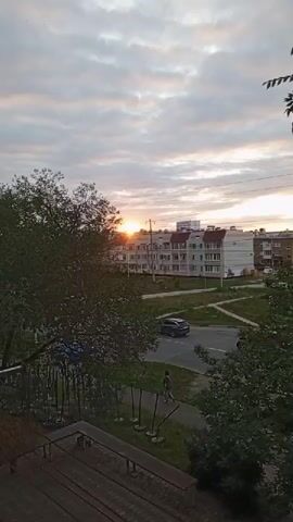 The video shows a view from a balcony overlooking a street and buildings at sunset. In the foreground, there are trees and part of a building structure. In the mid-ground, a street runs horizontally, with a few cars and a person walking. A dark SUV is seen driving on the street. A person on a bicycle is also visible. In the background, several multi-story buildings are present, with the setting sun partially visible between them, casting a warm glow on the clouds. The sky is cloudy, with streaks of orange and yellow indicating the sunset.