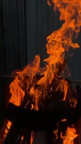 This video captures a close-up view of a bonfire at night. The flames are vibrant orange and yellow, dancing and flickering intensely. In the foreground, the dark, silhouetted rim of a fire pit is visible, with logs or burning material obscured within its base. The background is dark, suggesting the video was filmed after sunset, with a dark, textured surface, possibly a corrugated metal wall, visible. Sparks and embers occasionally rise from the flames, disappearing into the darkness. The overall impression is one of warmth and natural energy against a dark, ambient setting.