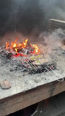 A grill rack with pieces of food, possibly fish, is placed over hot coals and an open fire. The fire is burning wood logs, producing flames and smoke. The grill rack is made of metal and has a handle. The fire is situated within a brick structure. The ground around the fire appears to be concrete or stone. The scene is outdoors, with a view of the sky in the background.