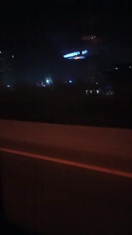 The video shows a view from a moving vehicle at night. The camera is positioned inside the vehicle, looking out the window. The scene outside the window is dark, with city lights visible in the distance. The lights include those from buildings, streetlights, and vehicle headlights. The vehicle appears to be traveling on a road or highway, with a low concrete barrier visible in the foreground. The overall impression is one of a nighttime urban landscape observed during travel.