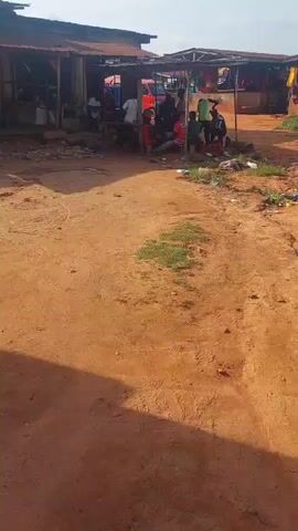 The video shows a group of people gathered under a makeshift shelter in an outdoor market area. The ground is dirt and sand, with some sparse vegetation and scattered litter. The shelter appears to be made of wood and corrugated metal, with several stalls or vendor areas visible. A red vehicle is parked behind the people. Several individuals are seated or standing, some appear to be engaged in conversation or activity, while others are simply present. The overall scene depicts a community gathering in a simple, rural or semi-urban environment.
