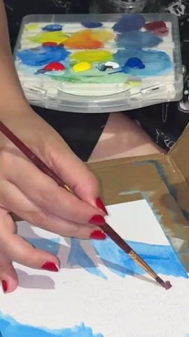 A close-up shot captures a person painting on paper with a thin paintbrush. The person's hands, adorned with red nail polish, delicately maneuver the brush, applying strokes of purple paint. Above the artwork, a clear plastic paint palette displays an array of colorful paints, including blue, red, yellow, and white. The background is blurred, hinting at the person's lap and patterned clothing.