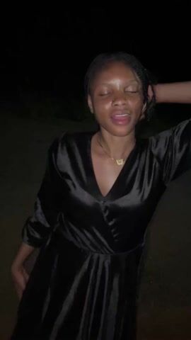 A young woman stands in a dark, low-lit outdoor area. She wears a shiny, black, V-neck dress with long sleeves and a gold necklace with a butterfly pendant. Her hair is styled in neat braids, and she has a nose piercing. The woman licks her lips and smiles, placing one hand on her head, then opens her mouth wide. The background is dark and blurred, suggesting it’s nighttime.
