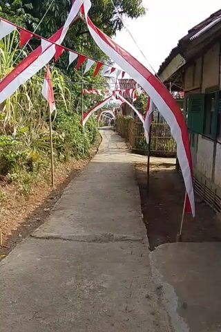 A paved pathway is lined with red and white Indonesian flags strung across it, forming arches. The pathway is flanked by lush green vegetation on the left and a rustic building with a thatched roof on the right. The scene suggests a festive or celebratory atmosphere.