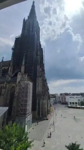 The video shows a view from a window overlooking a large Gothic cathedral, possibly undergoing renovation, as evidenced by the extensive scaffolding covering its exterior. The cathedral's tall spires dominate the skyline, with a bright, partly cloudy sky overhead suggesting it's daytime, likely late morning or early afternoon given the strong sunlight.

In the plaza in front of the cathedral, a few people are visible, some walking, others perhaps sitting. The plaza appears to be a public space, paved with light-colored stones. A few trees are also present, adding greenery to the scene. Modern buildings can be seen in the background, contrasting with the historical architecture of the cathedral.

The overall impression is one of a serene, urban environment on a pleasant day. The activity is minimal, suggesting a typical day in the city. No text is visible in the video.