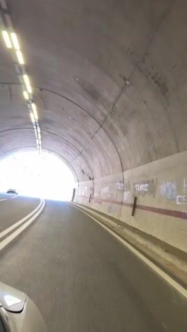 The video shows a first-person perspective of a vehicle traveling through a tunnel and then emerging onto a winding road with a scenic mountain view. The tunnel is made of concrete with overhead lighting and some graffiti on the walls. The road outside the tunnel is paved with white lines and has a guardrail along the side. The surrounding environment consists of a steep, rocky mountainside covered with vegetation and trees. The sky is blue with scattered clouds. In the distance, more mountains are visible. There are other vehicles on the road and a street lamp is visible.