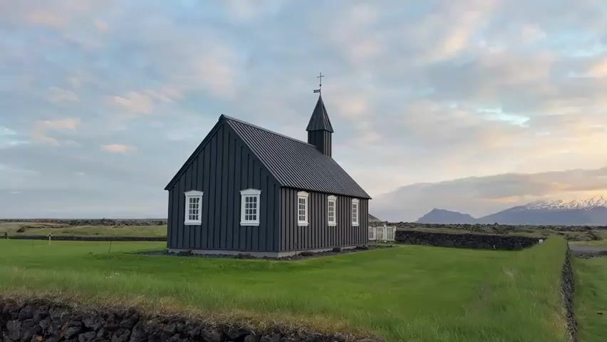 The video opens with a shot of a distinctive black wooden church with white-framed windows and a small steeple, positioned on a vibrant green lawn. A low, dark stone wall runs along the foreground. In the background, a dramatic landscape unfolds with dark, rugged mountains, some of which are partially covered in snow. The sky is filled with scattered clouds, illuminated by warm hues of orange, yellow, and pink, suggesting either sunrise or sunset.

As the camera slowly pans to the right, the church moves out of frame, revealing more of the vast, open scenery. A prominent, dark mountain dominates the mid-ground. The colorful sky continues to draw the eye, casting a soft, ethereal light over the entire scene. Green fields stretch across the landscape, with a narrow road or path visible in the distance. The stone wall extends along the right side of the frame, bordering the grassy area. The overall scene depicts a serene and picturesque natural environment. No people or visible text are present.