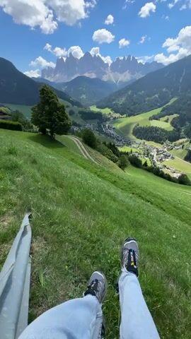 The video shows a first-person view of someone sitting on a grassy hillside, with their legs and feet in the foreground. The person is wearing blue jeans and athletic shoes. Behind them, a valley unfolds, dotted with houses and trees, and further in the distance, a majestic mountain range rises under a bright blue sky with scattered clouds. A metal object, possibly part of a bench or railing, is visible on the left side of the frame. The scene appears to be a scenic overlook in a mountainous region.